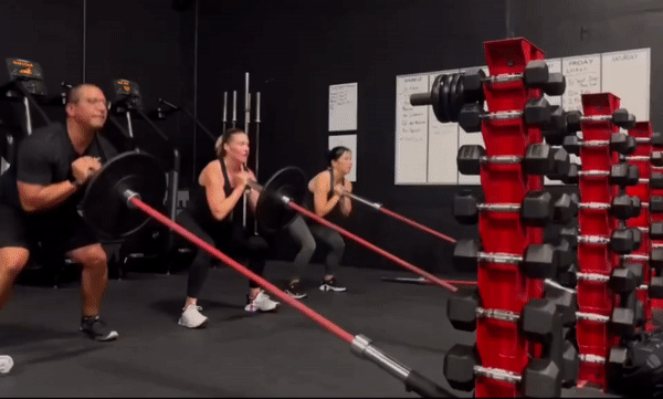 a short video of a group of people doing landmine workouts using Synergy Custom Fitness's landmine attachment accessory at the base of several red vertical dumbbell racks in a commercial gym.
