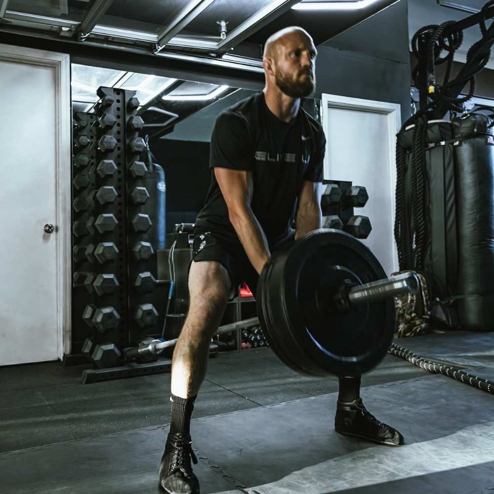 a man doing squats with a barbell connected to a black 10-tier vertical dumbbell rack by a landmine attachment accessory.