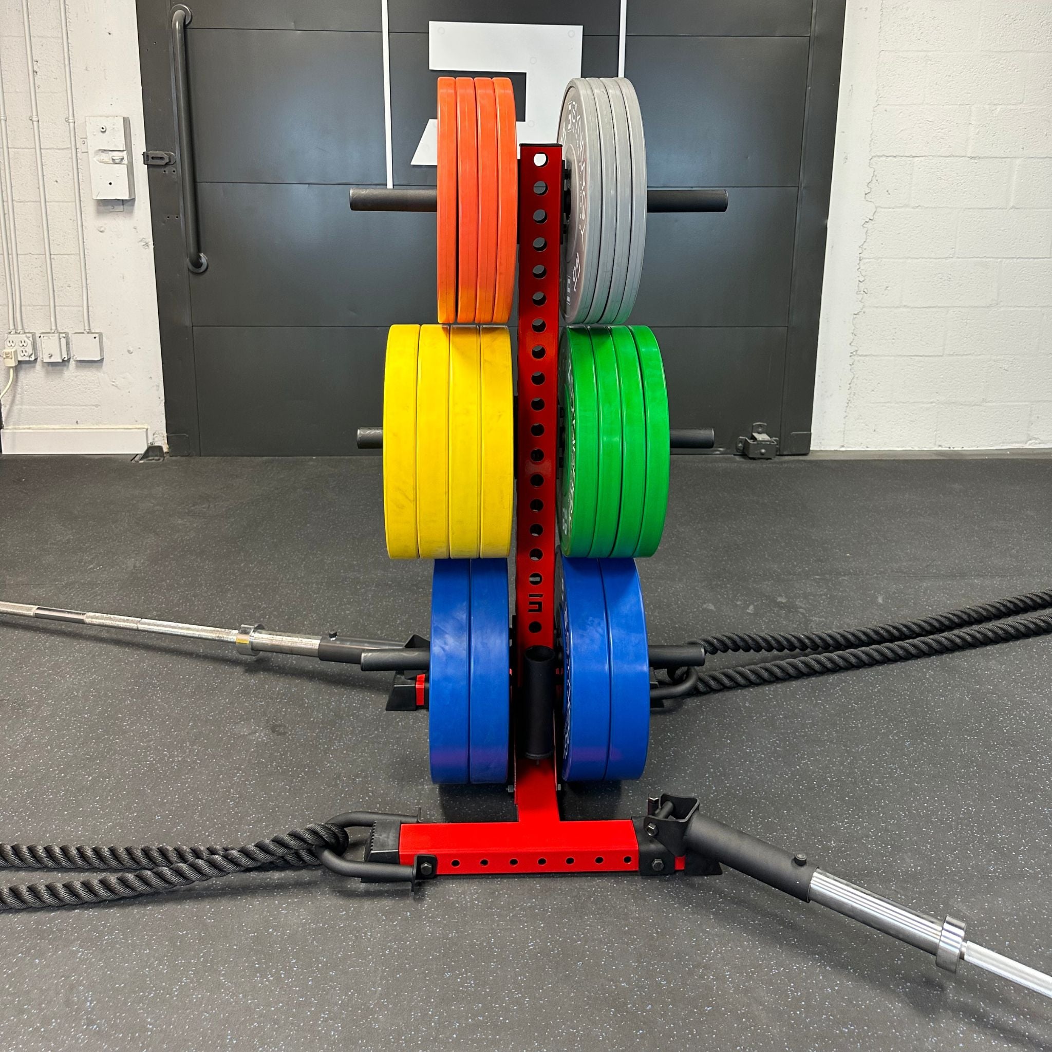 red alpha vertical weight plate storage tree with six weight plate holders holding various weight plates, two barbells, and two battle ropes using battle rope and landmine accessory attachments