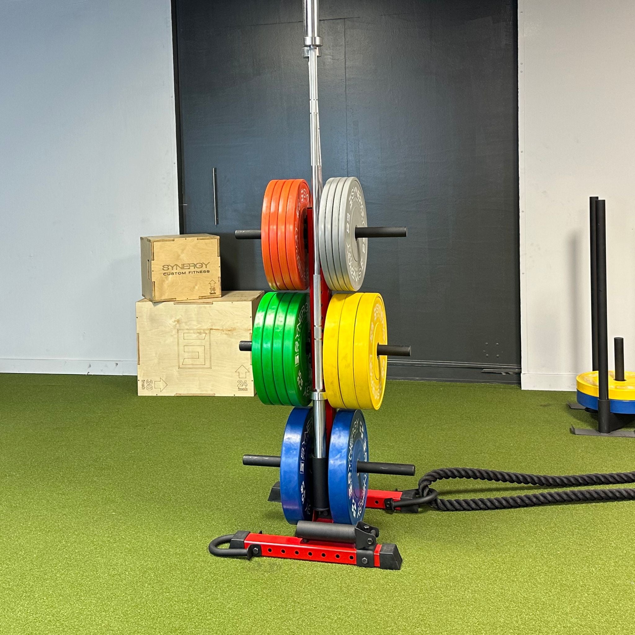 red vertical weight plate storage tree with six weight plate holders holding various weight plates, a barbell, and a battle rope using a barbell holder and battle rope accessory attachment