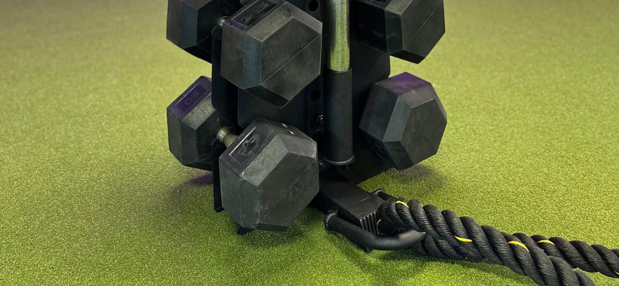 the base of a black vertical dumbbell rack holding dumbbells, a barbell being help by a barbell holder accessory, and a battle rope being held by a battle rope accessory.