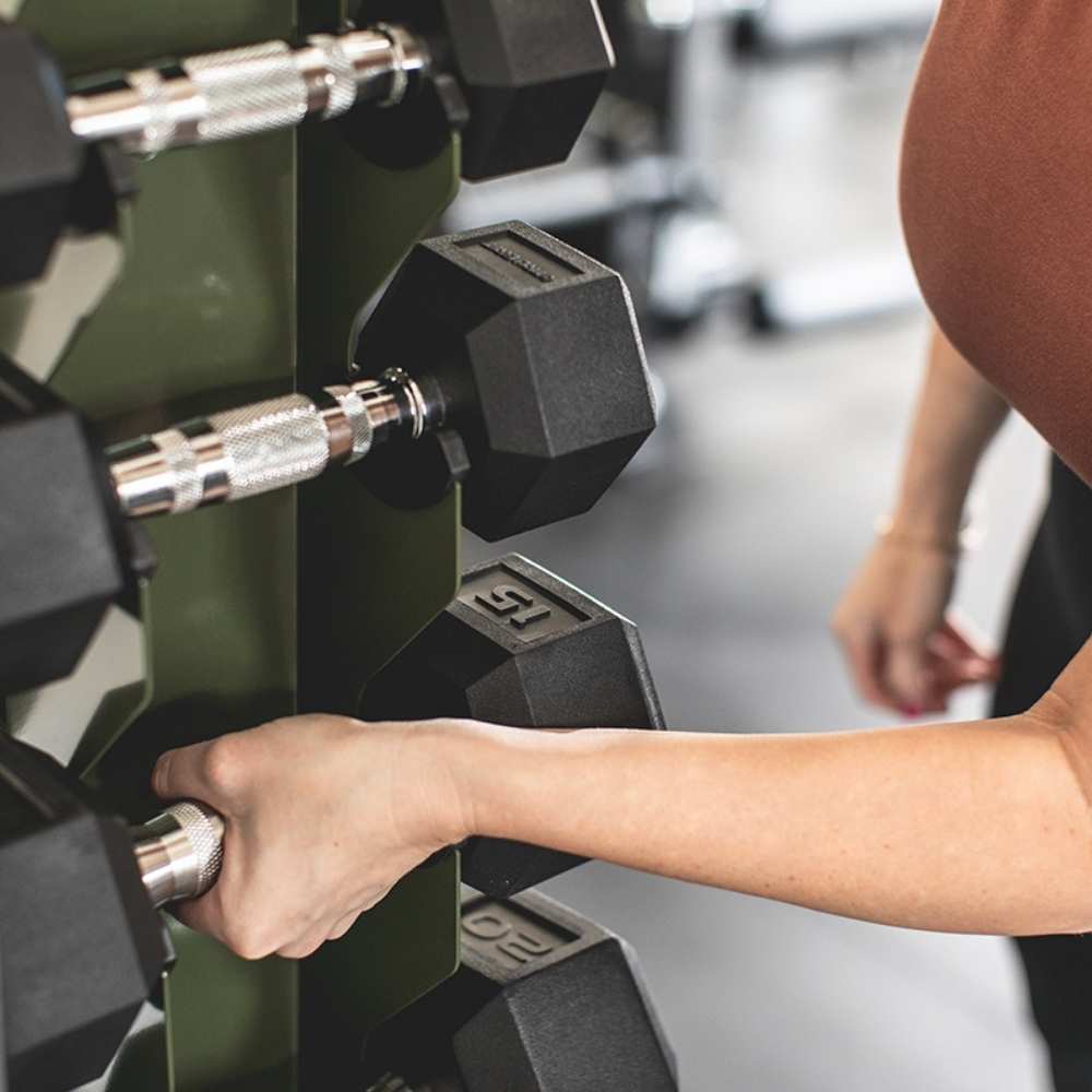 a woman taking a dumbbell off a Synergy Custom Fitness military green vertical dumbbell rack.