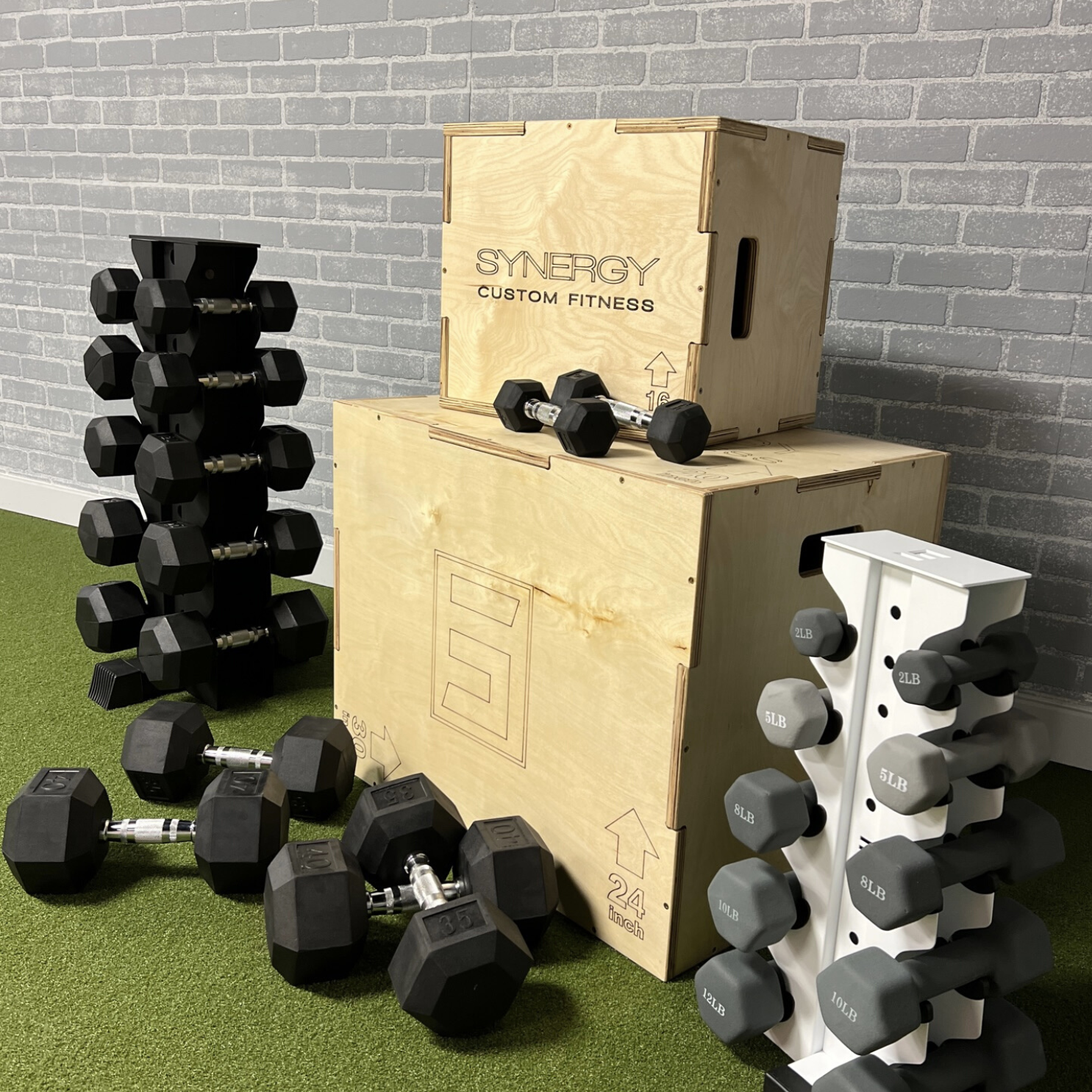 two wooden plyo boxes with the smaller plyo box stacked on a larger plyo box surrounded by dumbbells, a black vertical dumbbell rack, and a white vertical dumbbell rack both loaded with dumbbells